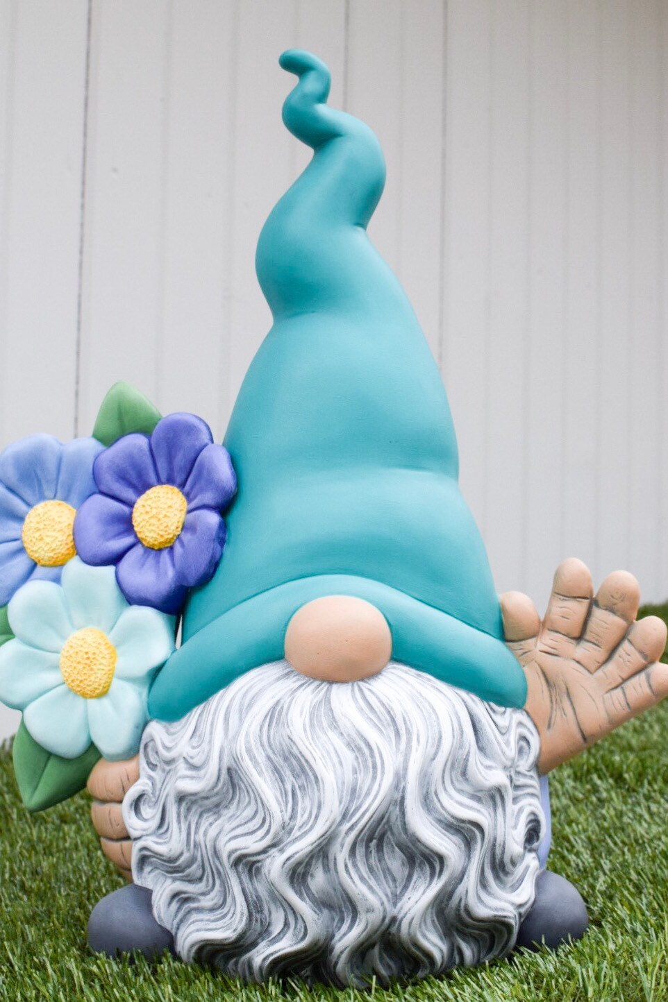 May include: A whimsical garden gnome figurine with a teal hat, gray wavy beard, and light blue flowers. The gnome's right hand is raised in a friendly wave.  The figurine is made of painted ceramic and has a charming, handcrafted look. It's a perfect decorative piece for gardens or homes.