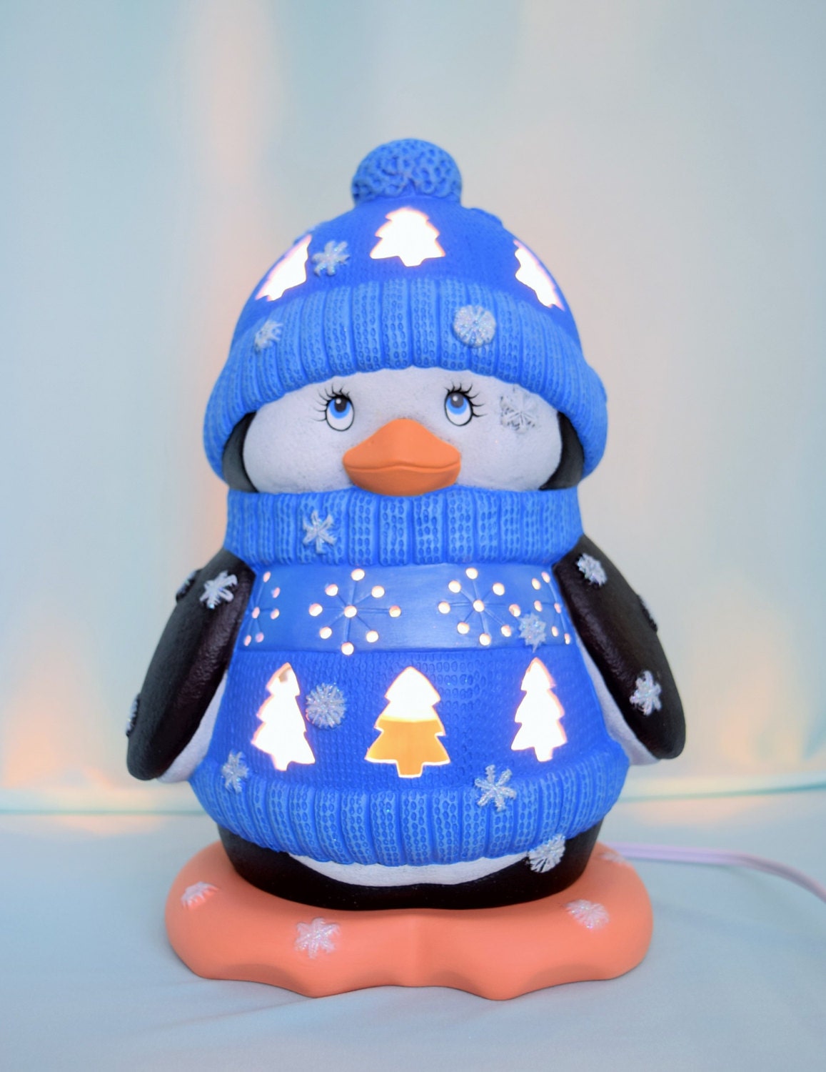 May include: A ceramic penguin figurine wearing a blue knitted hat and sweater with snowflake and tree designs. The penguin has a white body, black wings, and a bright orange beak. The figurine is lit from within, creating a warm glow.