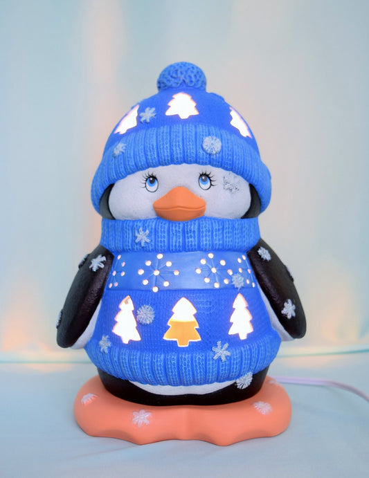 May include: A ceramic penguin figurine wearing a blue knitted hat and sweater with snowflake and tree designs. The penguin has a white body, black wings, and a bright orange beak. The figurine is lit from within, creating a warm glow.